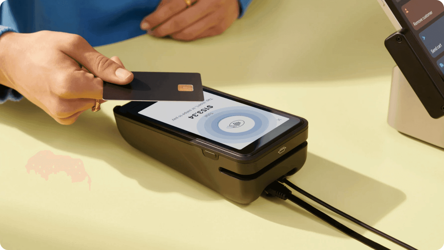 A customer holding a bank card over a countertop payment terminal showing a $153.34 contactless prompt